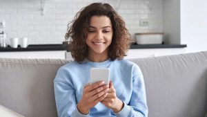 Mulher sentada no sofá segurando um celular. Foto: Adobe Stock