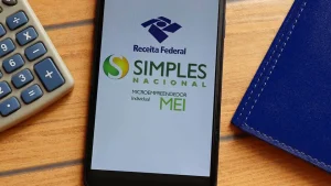 Celular apresenta logo da Receita e do Simples Nacional e MEI, juntamente com calculadora e caderno. Ferramentas que auxiliam no cálculo do IR. Foto: Brenda Blossom - Adobe Stock