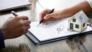 Pessoa assinando contrato de compra com um miniatura de casa sobre os papéis