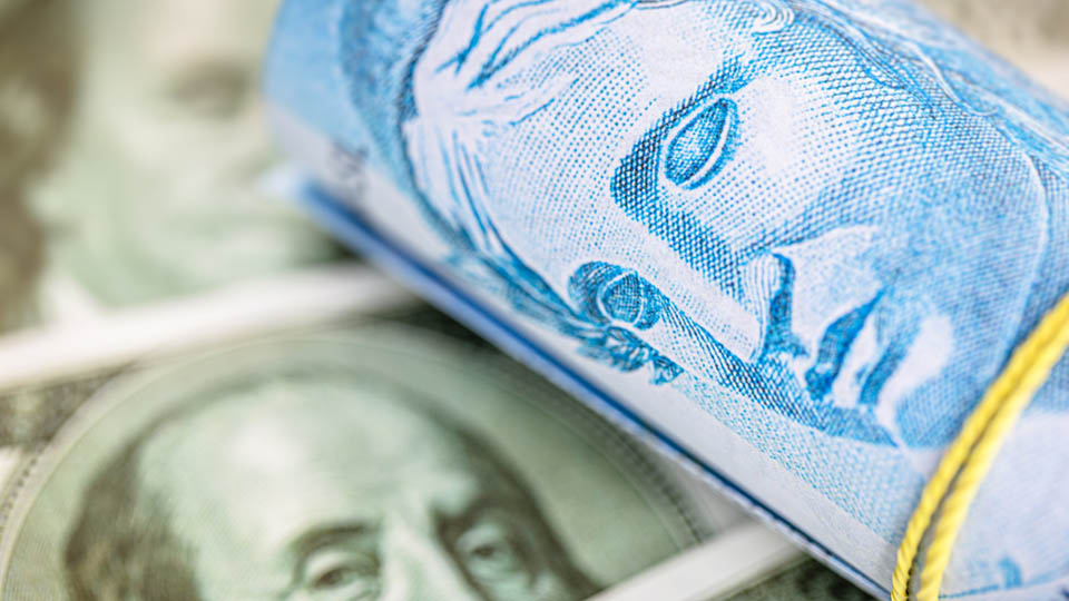 Rolo de cédulas de Real cruzada sobre notas de Dólar. Foto: Adobe Stock