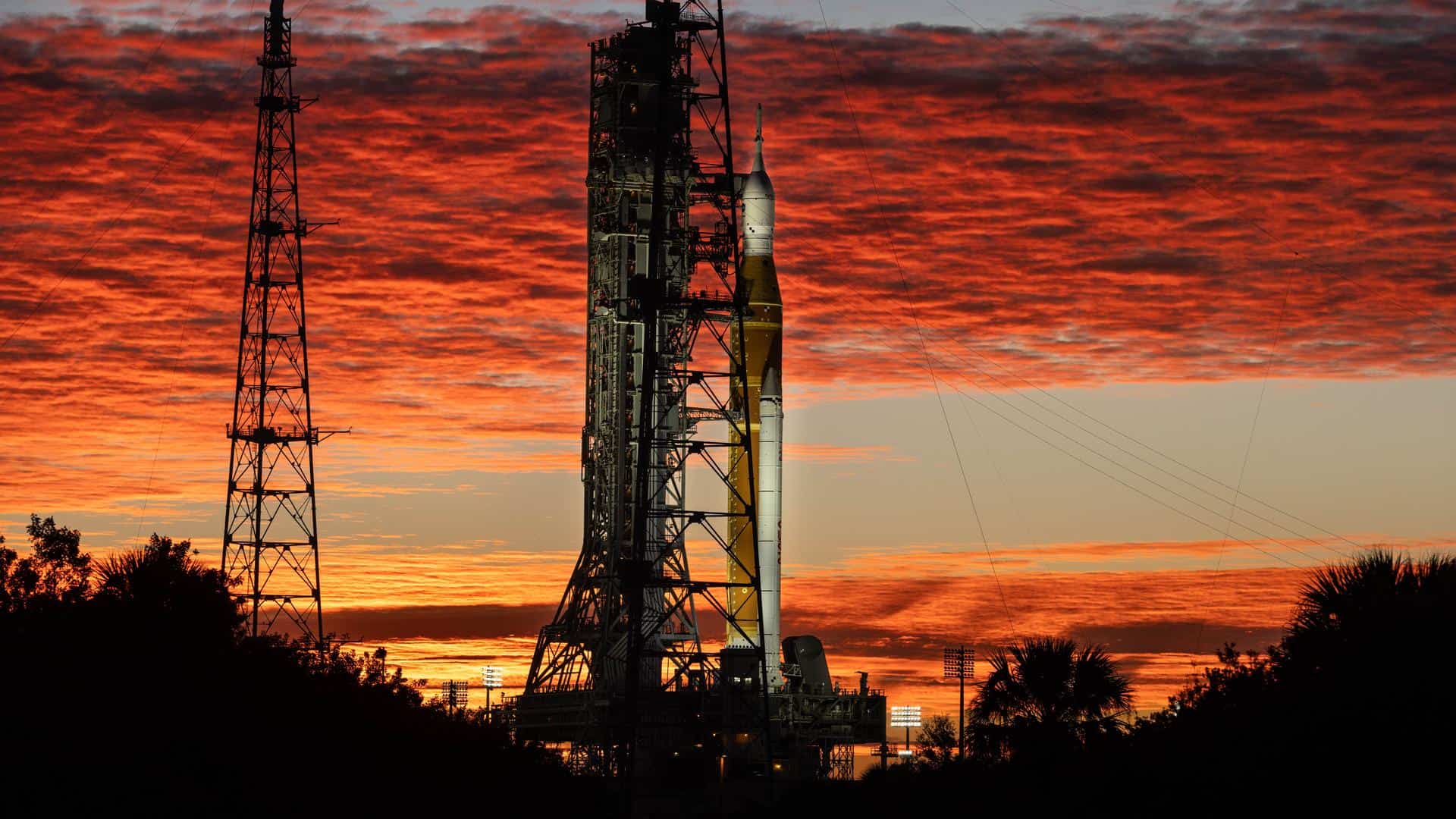Silhueta do foguete SLS e da nave Orion, da missão Artemis II, na Plataforma de Lançamento 39B do Kennedy Space Center na Flórida. O céu está dramaticamente iluminado com nuvens em tons de vermelho e laranja pelo nascer do sol. Árvores e torres de transmissão aparecem em silhueta no primeiro plano
